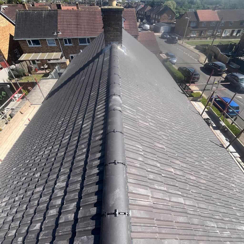 View along a grey tiled roof ridge with mortared ridge tiles and a brick chimney, overlooking a residential street.