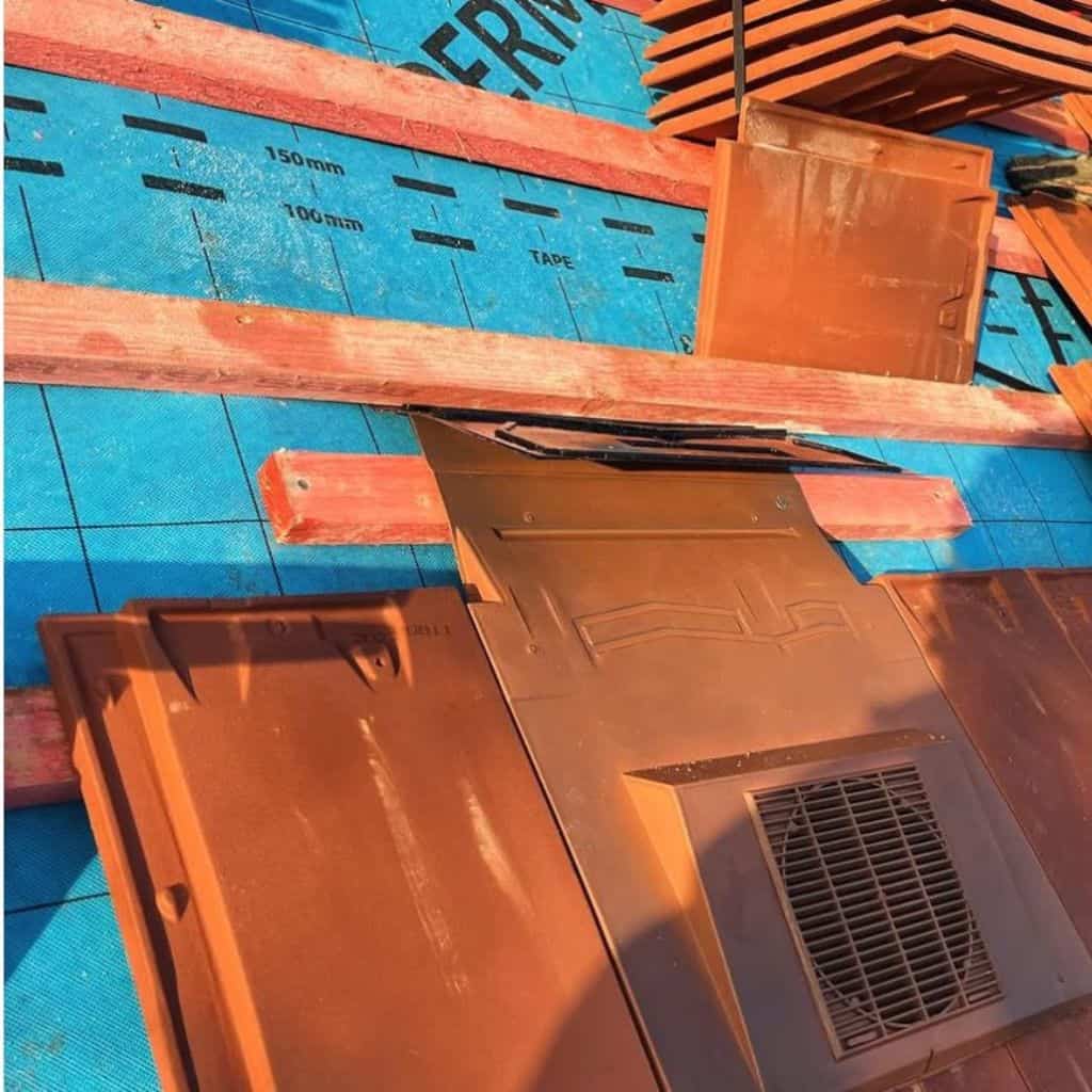 Red clay roof tiles and a tile vent being laid over blue breathable underlay and timber battens in sunshine.