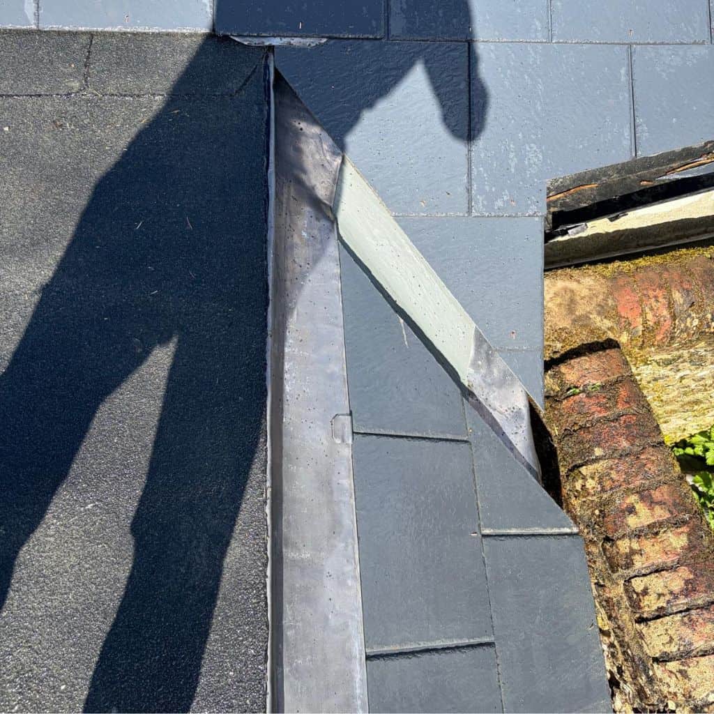 Close detail of grey lead flashing dressed into a valley between two slate roof slopes, with old brickwork to the right.