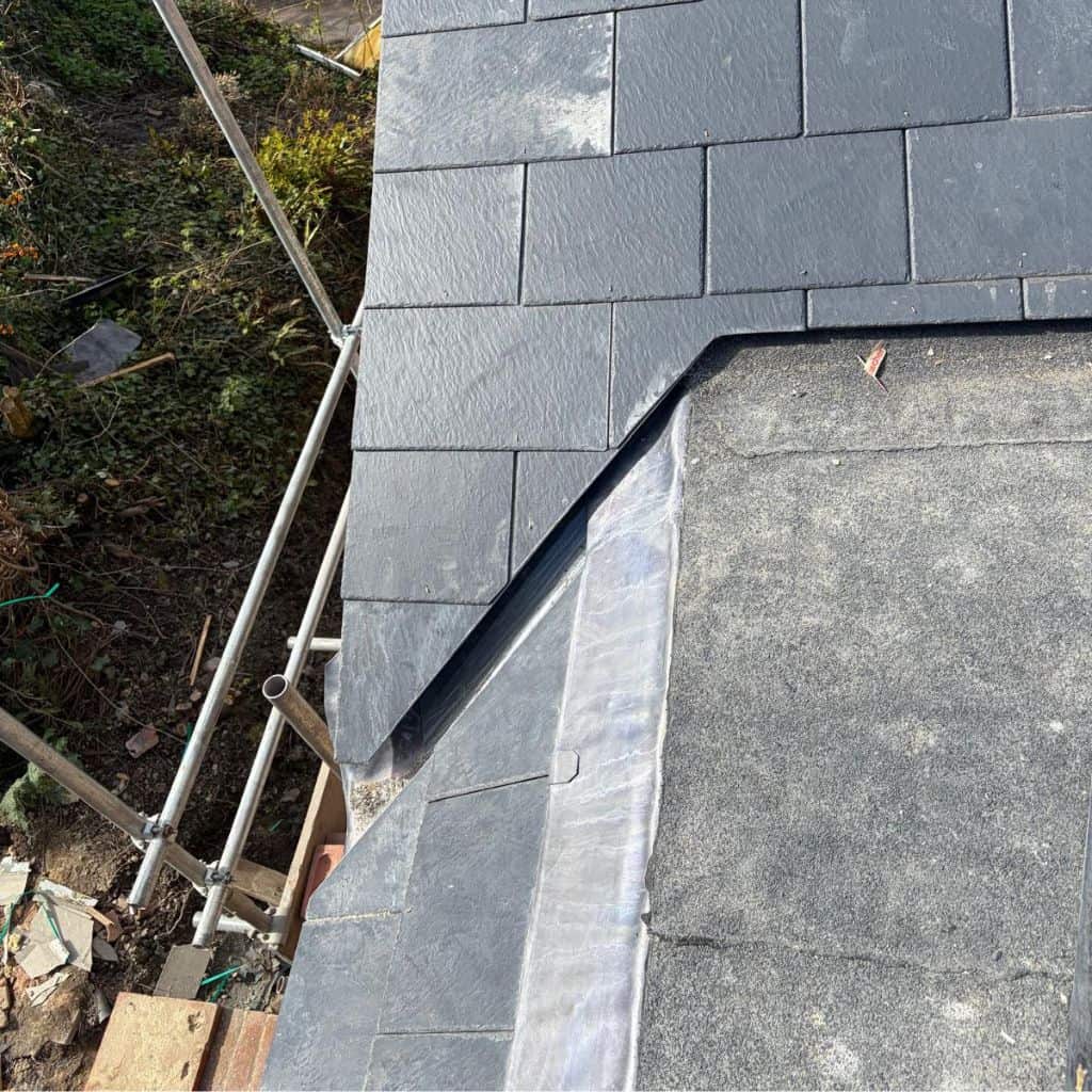 Grey lead-lined roof valley running between dark slate tiles and a felt flat roof section, with scaffolding to the side.