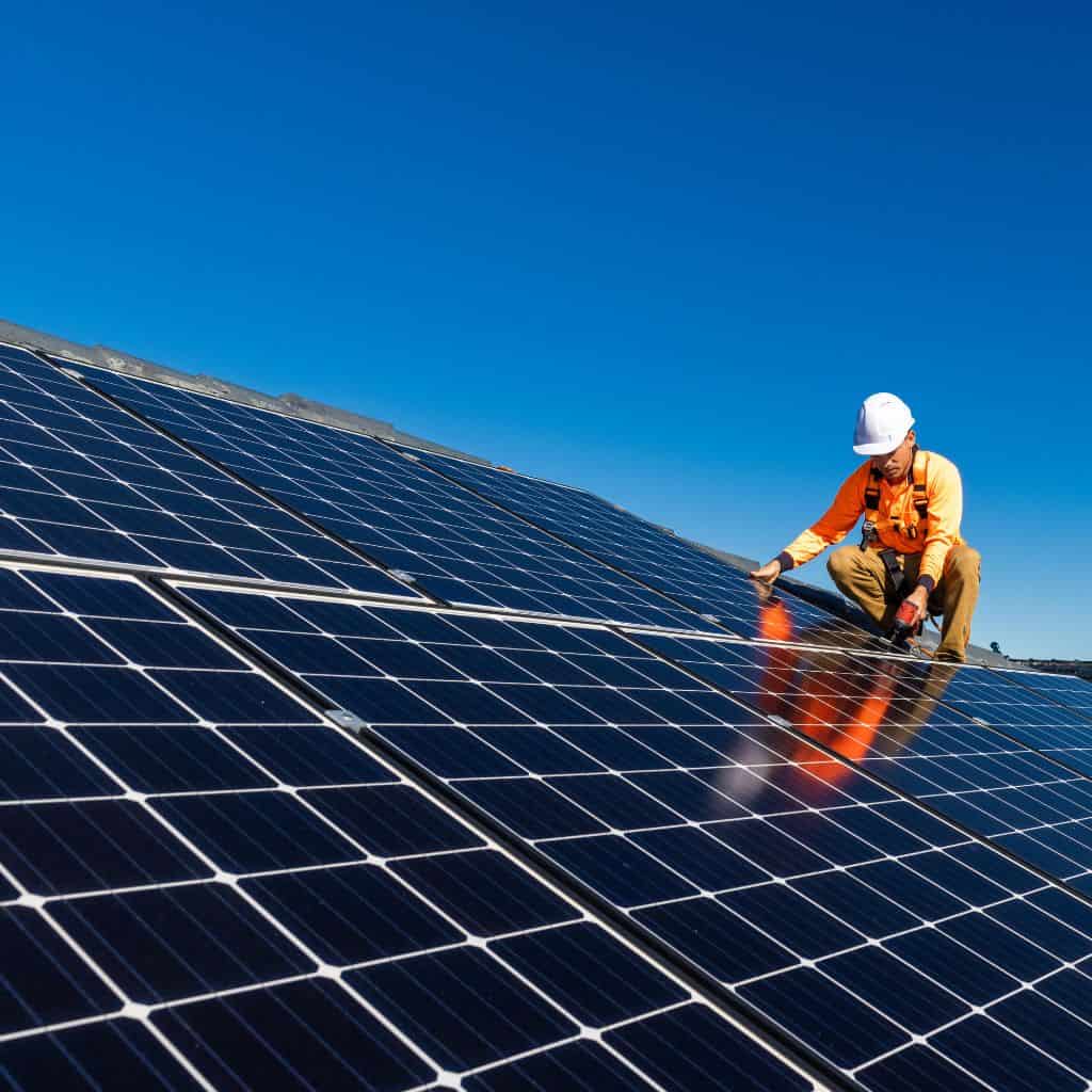 Tradesman in an orange hi-vis shirt and hard hat kneeling beside a large array of dark rooftop solar panels under blue sky.
