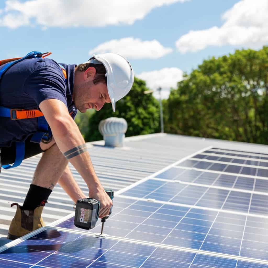 Installer in a hard hat and harness kneeling to fix a solar panel to a rooftop with a cordless drill, green trees behind.