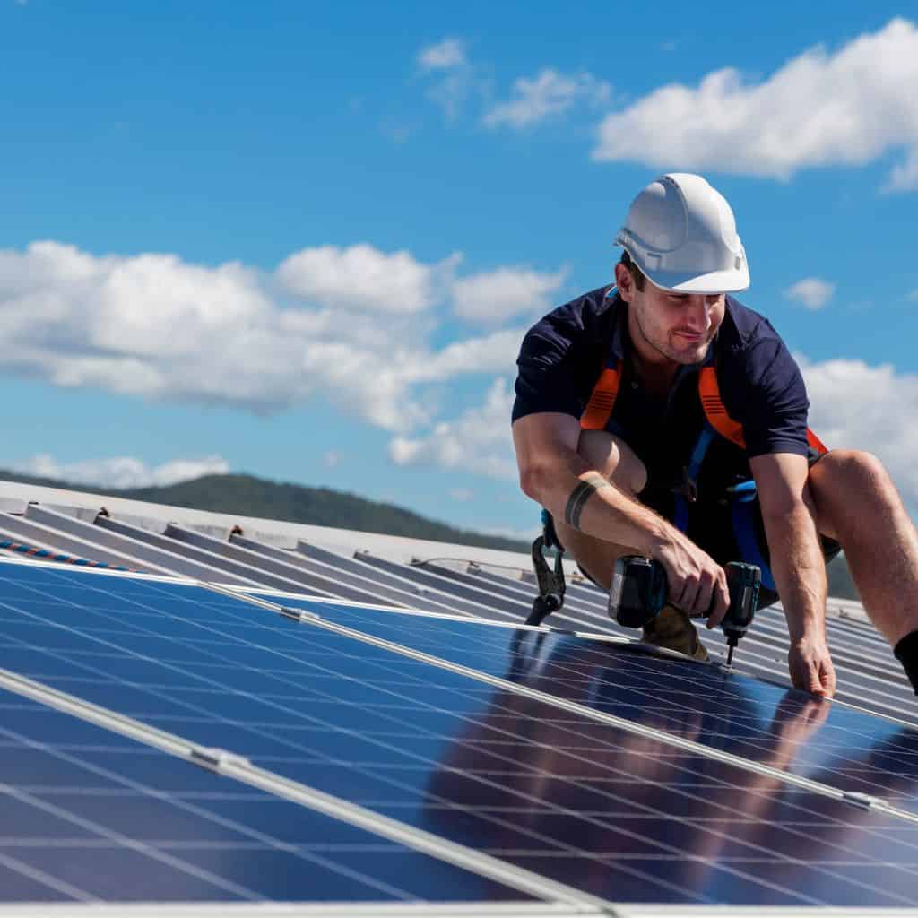 Installer in a hard hat and harness drilling a solar panel into place on a sunny rooftop array.