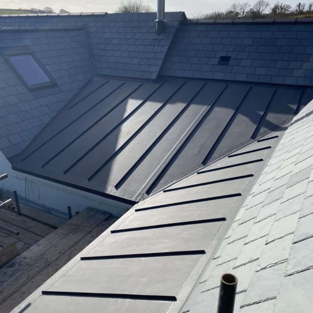 Grey standing-seam single-ply membrane roof meeting slate-tiled pitches, with a flue and rooflight nearby.