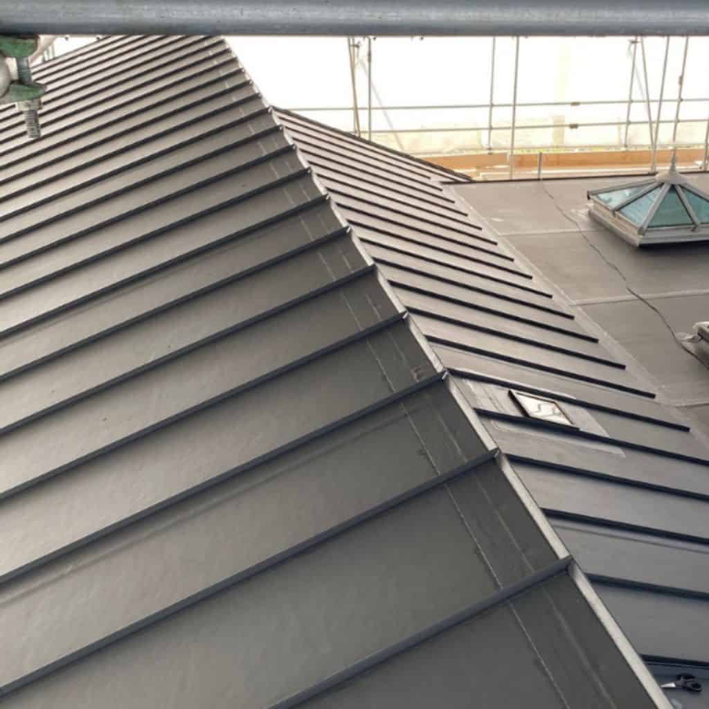 Close view of a grey standing-seam metal roof meeting a flat roof section with a glazed lantern, framed by scaffolding.