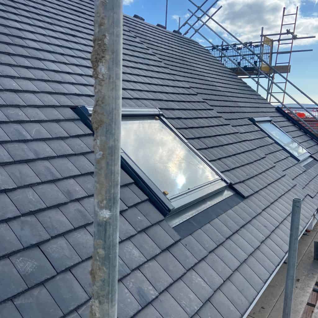 Grey slate-tiled pitched roof with a flush-fitted skylight window, scaffolding and a blue sky in the background.