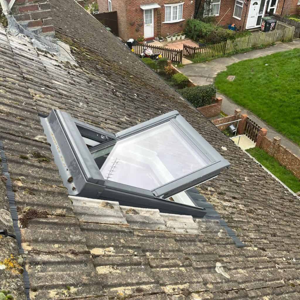 Grey-framed roof window propped open on a weathered tiled roof, overlooking brick houses and a front garden.
