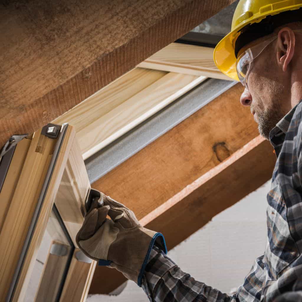 Worker in a yellow hard hat and safety glasses fitting a timber-framed roof window from inside a loft.