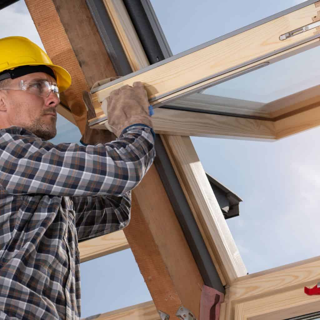 Worker in a yellow hard hat and safety glasses fitting a timber-framed roof window from inside against the sky.