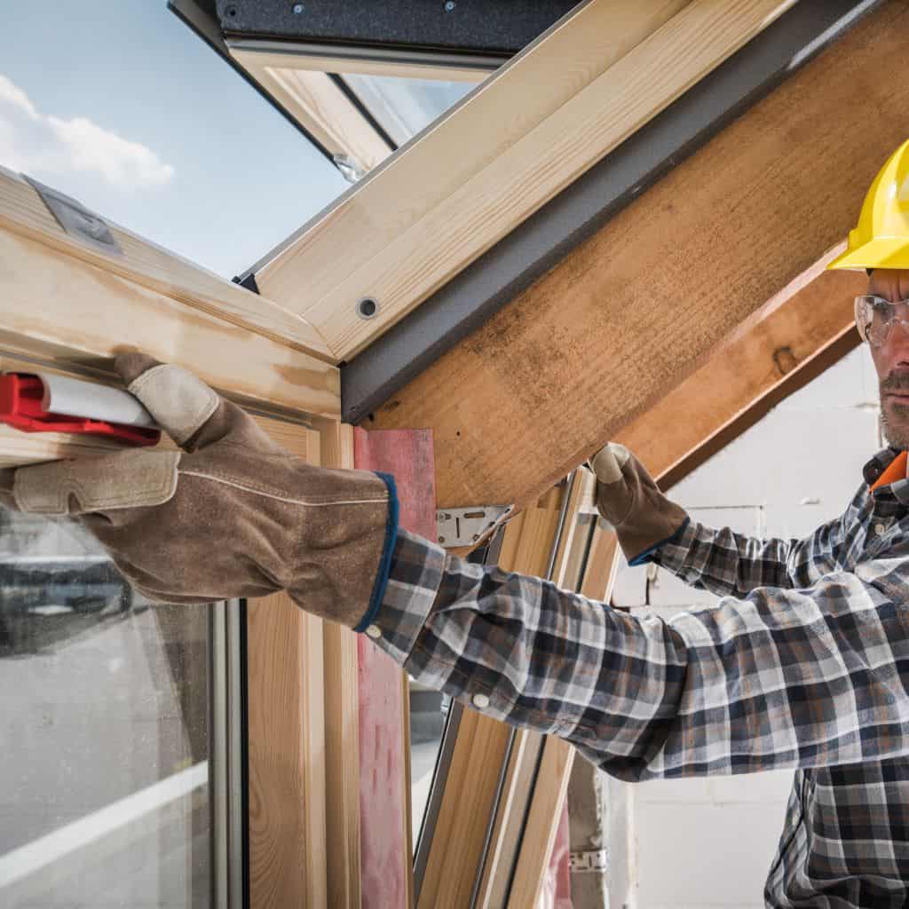 Roofer in a yellow hard hat and gloves fitting timber-framed roof windows from inside a pitched roof opening.