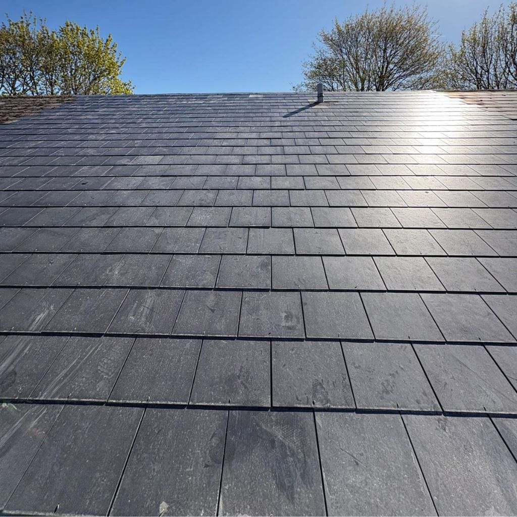 Expanse of newly laid dark grey slate roof tiles in neat overlapping rows, sunlight catching the upper slope under blue sky.