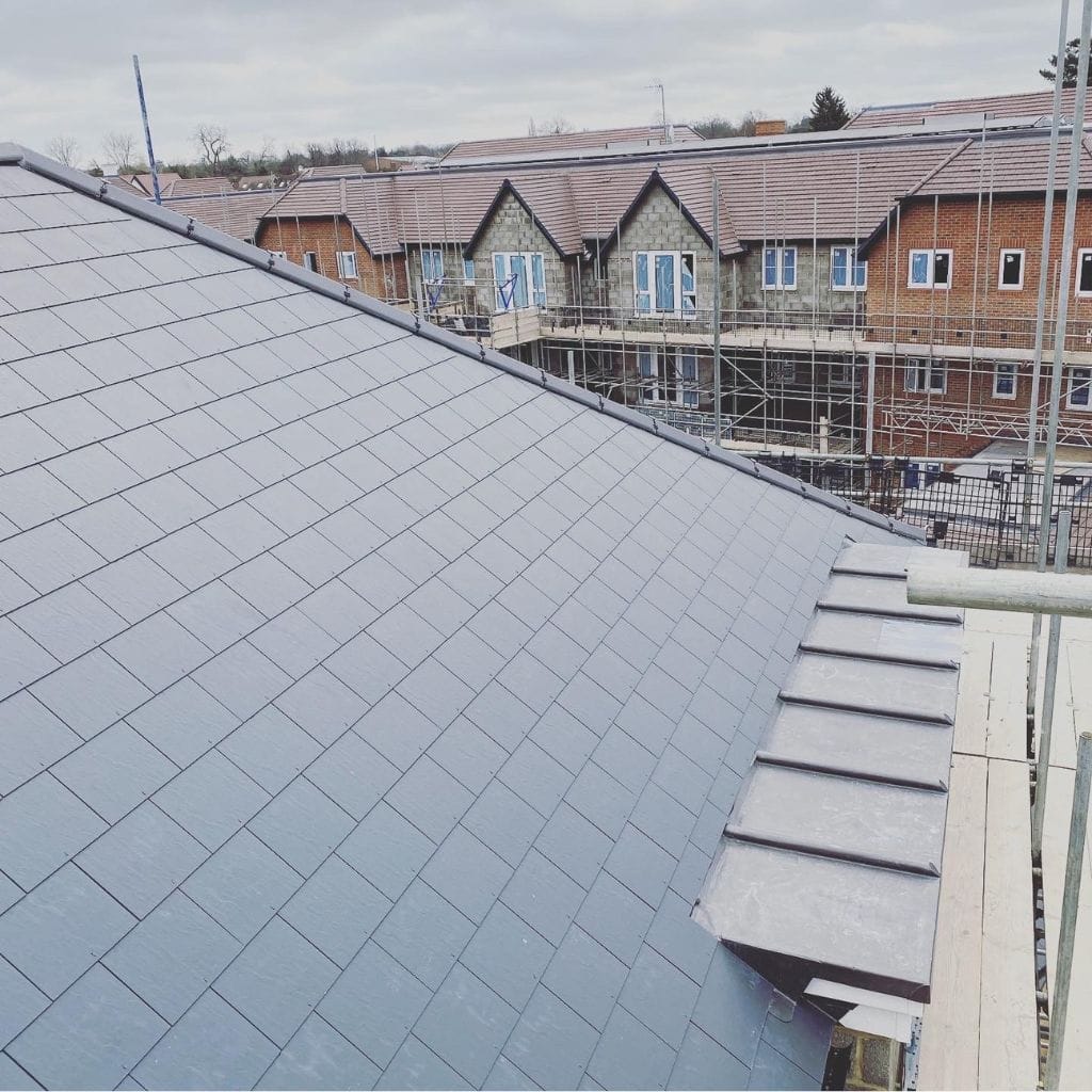 Expanse of grey slate roof tiles on a new build, with a leadwork hip and other houses under construction behind.