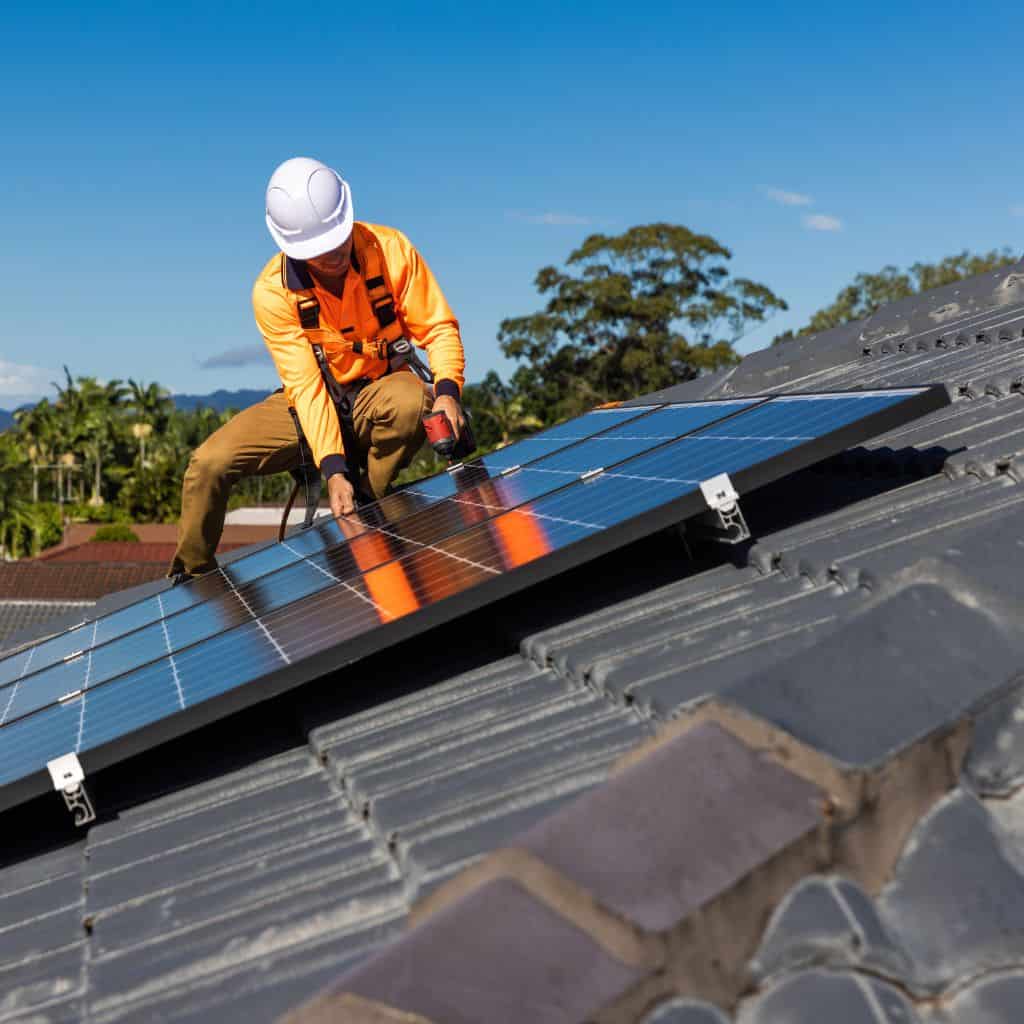 Worker in an orange shirt and white hard hat drilling solar panels onto a grey tiled roof under a clear blue sky.