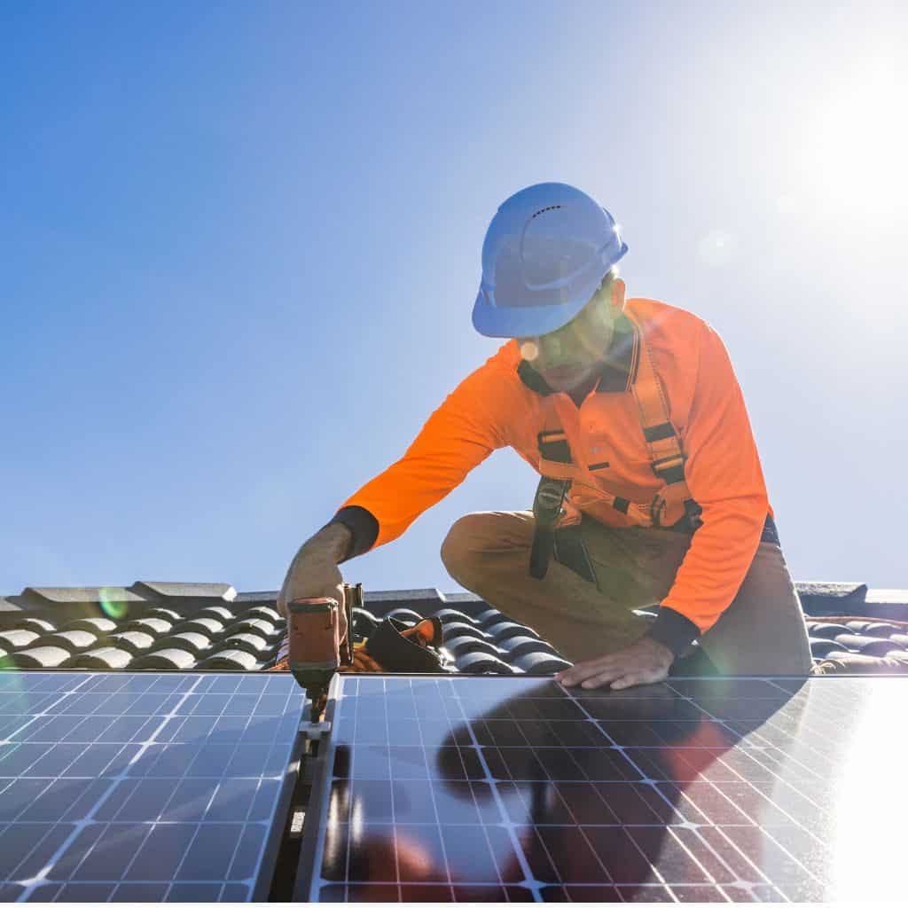 Worker in an orange shirt, hard hat and harness drilling a solar panel into a mounting rail on a tiled roof.