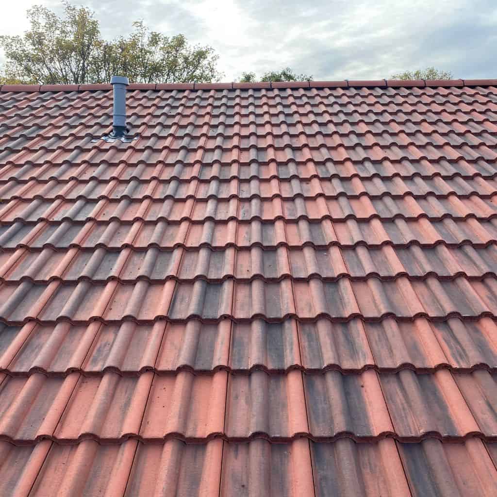 Expanse of red and brown interlocking pantile roof with a grey flue vent near the ridge, trees and sky behind.