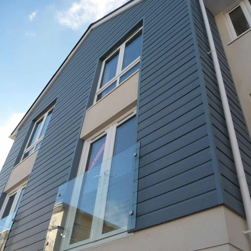 Building corner clad in blue-grey horizontal weatherboard with white uPVC windows and a glass balustrade.