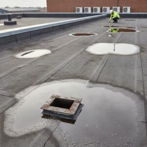 Worker inspecting flat roof with multiple waterproofing patches and drains.